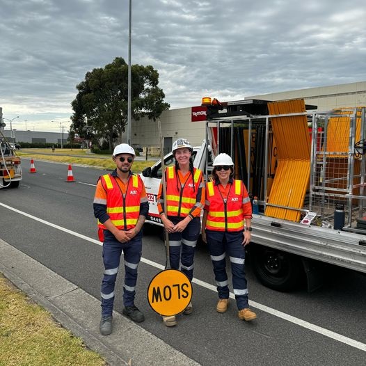 Three A2Z Traffic workers stand by a traffic control vehicle, holding a "SLOW" sign, ready for roadwork operations. Ask ChatGPT