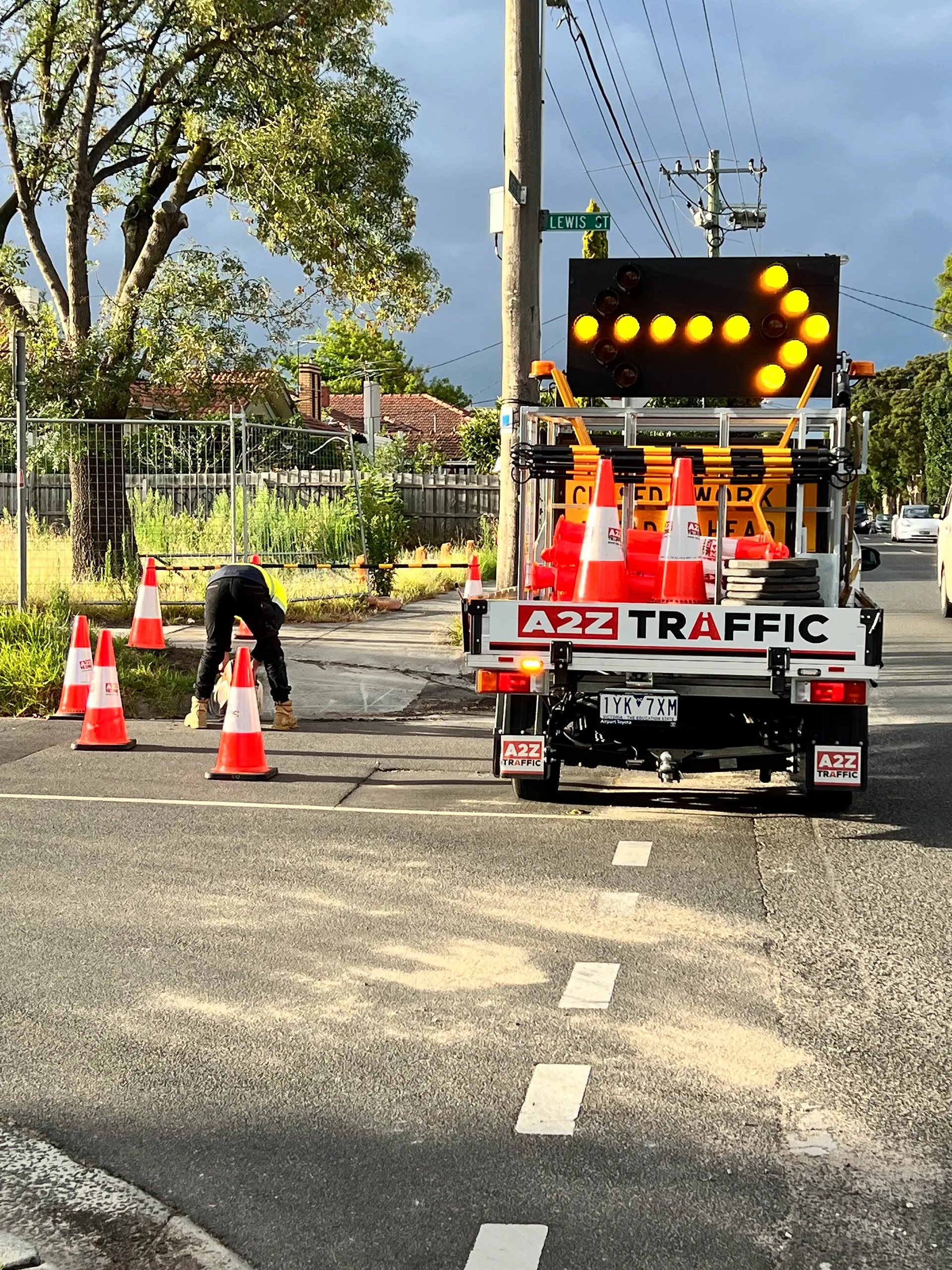 A2Z Traffic ute with a flashing right-arrow board while a worker sets out cones at a suburban roadwork site.