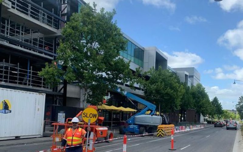 A2Z Traffic controller managing lane closure with a "SLOW" sign on a city street, ensuring safety for construction workers operating elevated equipment near a commercial building in Melbourne.