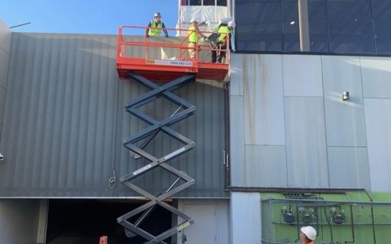 A2Z Traffic team member monitoring safety while workers operate a scissor lift on a commercial building site, with traffic cones and caution tape securing the area for pedestrian and vehicle protection.