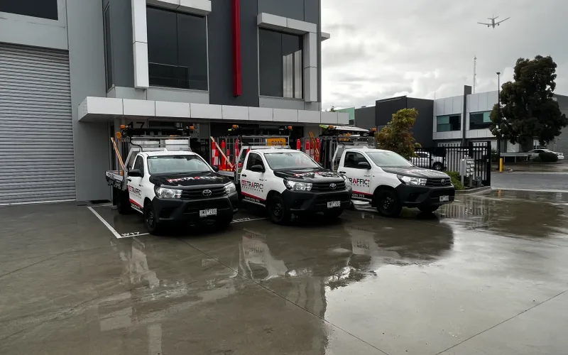Three A2Z Traffic utility utes parked on a wet forecourt outside an industrial building on a rainy day, with a plane visible in the cloudy sky.