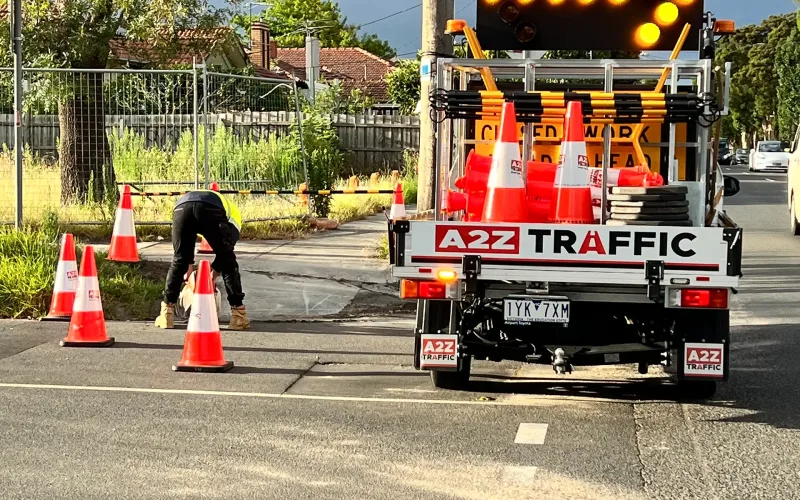 A2Z Traffic ute with a flashing right-arrow board while a worker sets out cones at a suburban roadwork site.